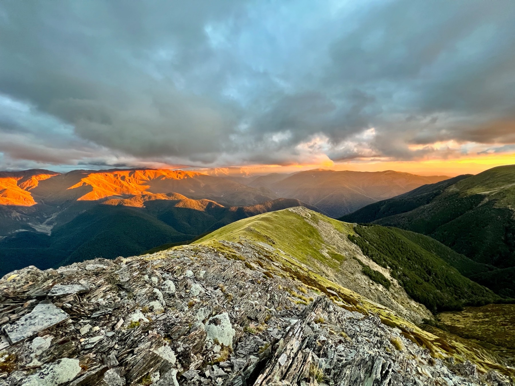Evening light on the far ranges and a rocky ridge in the foreground, under a heavy grey sky.